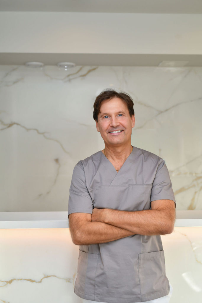 Smilende mann i grått helseantrekk med armene i kors foran marmormønstret vegg – representerer profesjonalitet og trygghet hos Drammen Tannlegesenter.