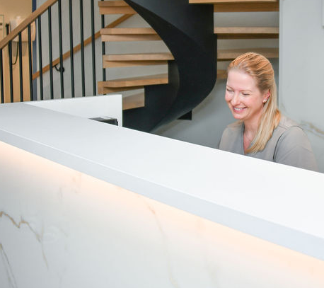Smilende resepsjonist bak moderne marmorskranke i Drammen Tannlegesenter, med spiraltrapp i bakgrunnen – et innbydende og profesjonelt miljø